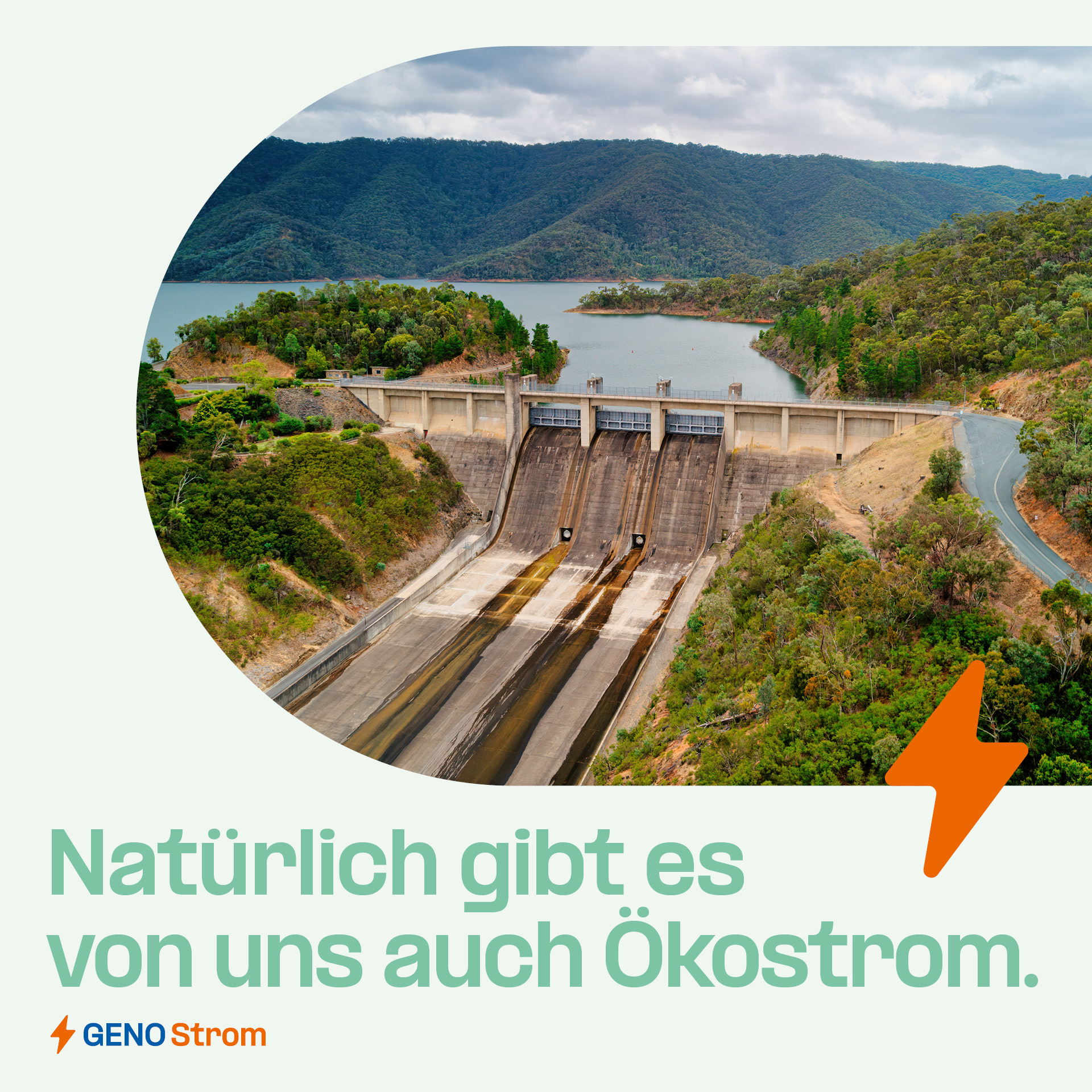 Eine Luftaufnahme eines großen Staudamms, umgeben von einer üppig grünen Landschaft mit Hügeln und einem Wasserreservoir im Hintergrund. Im Bild steht der Text: "Natürlich gibt es von uns auch Ökostrom. GENO Strom".